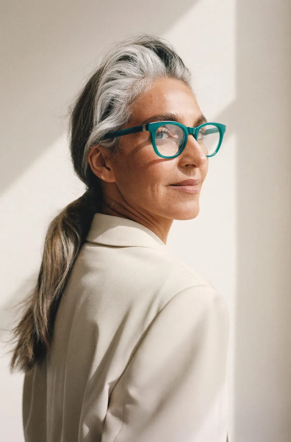 A woman with silver hair wearing bold teal glasses, looking back over her shoulder in soft window light
